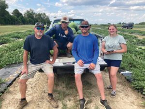 Three men sitting in the bed of a pickup truck, and one woman standing next to the truck in a field