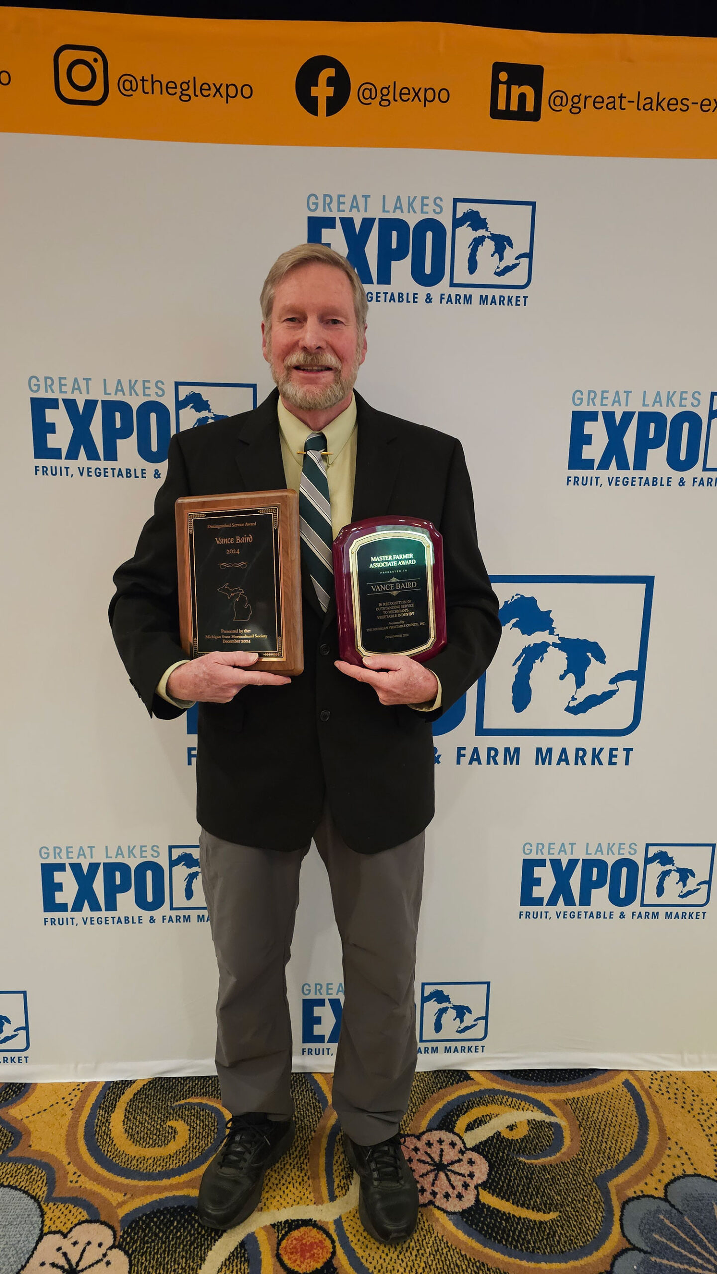 Man in a dark sport coat holding two plaques in from of a Great Lakes EXPO banner