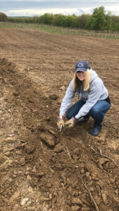 Blonde-haired woman in a baseball cap kneeling in a field with a tool in her hand