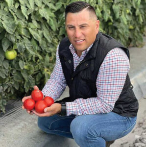 A dark-haired man crouched down holding four tomatoes in his hands