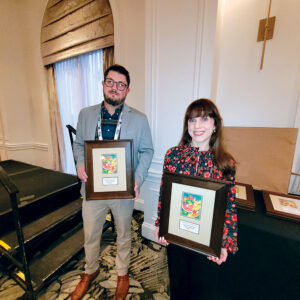 Man in gray sport coat, left, and a woman in a floral blouse, right, pose with framed awards
