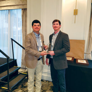Man in tan sport coat, left, handing glass award to a man in a dark gray sport coat, right